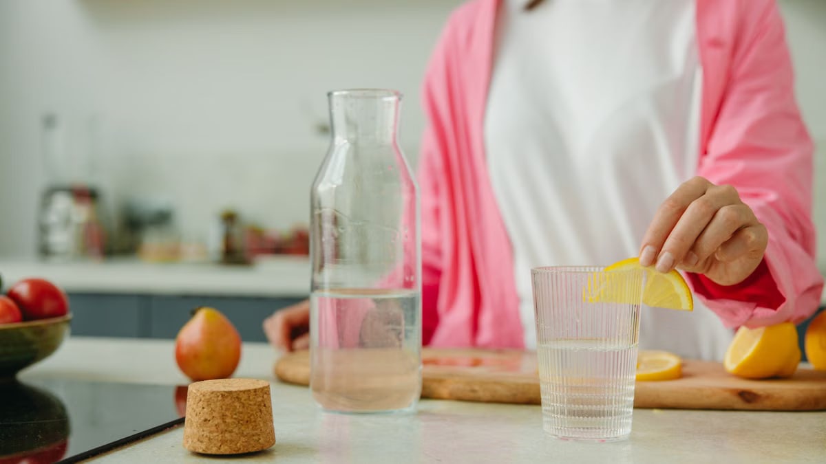 A woman in a pink outfit prepares lemon water in a bright, modern kitchen setting.